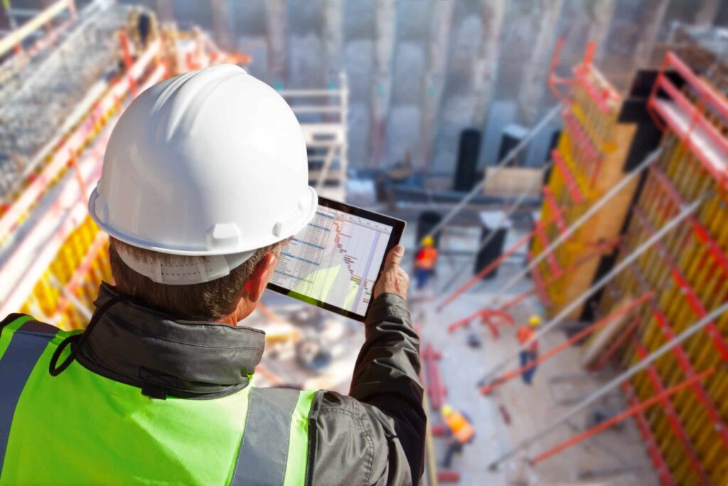 A mechanical contractor examining a roof installation plan on Jonas Construction Software via a tablet, strolling on a high-rise construction site with many other specialty contractors and heavy machinery in operation.