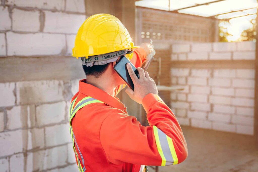 Jonas Construction worker in safety gear on a phone call at a construction site against the backdrop of a sunset, indicating round-the-clock services and software solutions. A partially built wall suggests ongoing construction project management.