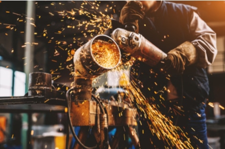 A construction worker wearing personal protective equipment skillfully operates an angle grinder on a metal piece, generating a bright cascade of orange sparks. The out-of-focus backdrop exudes the dynamic and high-temperature conditions inherent in metalworking tasks. This scene unfolds inside a Jonas Construction workshop - reflecting the practical, hands-on impact of our integrated software solutions for mechanical and specialty contractors.