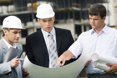 Three construction professionals reviewing a large blueprint at a job site, with the central figure in business attire pointing out a particular area of interest.