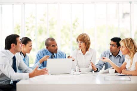 A group of diverse businessmen and businesswomen engaging in a lively discussion around a modern office table, with one individual accessing Jonas Construction Software on a laptop.