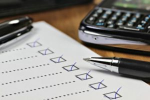 Alt text: A close-up image of a checklist on a paper with items marked off in blue ink, with a pen resting on top. In the background, a calculator and smartphone are partially visible on a wooden desk, indicating the integration of traditional and digital tools in project management.