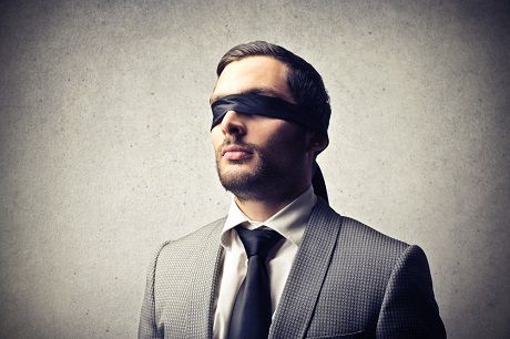 Alt: "Bearded man in a light gray suit, blindfolded with a black cloth, standing contemplatively against a textured gray background.