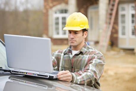 Construction worker in safety gear using Jonas Construction Software on a laptop for Accounting & Payroll, placed on the hood of a vehicle at a construction site with an under-construction house visible in the background.