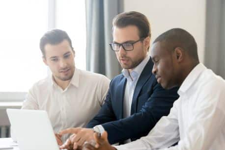 Three professional men, identified as specialty contractors, are engrossed in reviewing content on a laptop in an office setting. They exhibit signs of engagement and collaboration. Two of the men are Caucasian while the other is African American. All three are dressed in formal business attire, emphasizing their professional roles within the construction industry.