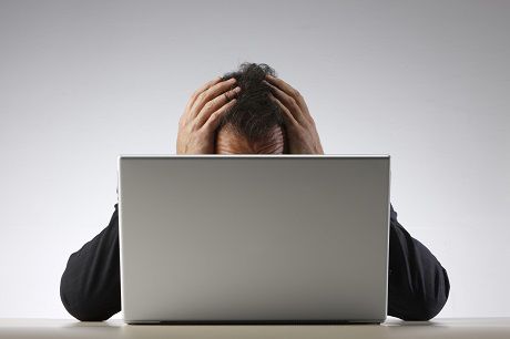 Alt text: An older man displaying signs of stress or confusion with his hands clutching his hair, sitting behind a laptop displaying Jonas Construction Software, placed against a light grey background.