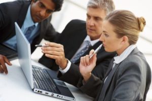 look on attentively from either side. One man is pointing to a particular feature of the software on the screen while the other has his arms crossed, denoting concentration and control. The scene depicts active discussion about construction project planning and management using Jonas Construction Software.