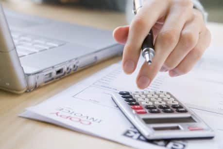 Hand calculating numbers with a pen and calculator, working on a laptop displaying Jonas Construction Accounting Software, surrounded by financial documents containing charts and graphs related to a construction project.