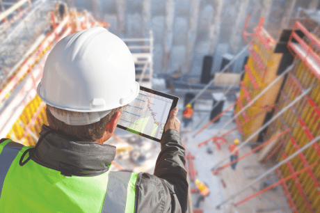 Construction professional reviewing data on a digital tablet amidst a busy work site, orchestrated by the sophisticated Jonas construction software, highlighted by elements like crane machinery and ongoing construction in the background.