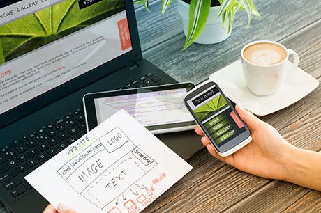 Alt text: A close-up shot of a hands holding a smartphone displaying a wireframe sketch. Wood desk background features essential workspace items including laptop, tablet and coffee cup - representing the technical work environment of specialty contractors utilizing Jonas Construction Software.