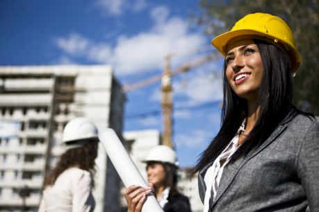 Alt text: Smiling female contractor in yellow hard hat and gray suit utilizing Jonas Construction software on construction site, holding blueprints with a crane, building under development and two colleagues in the background.
