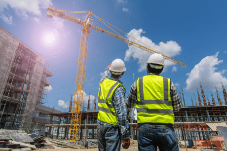 Two mechanical contractors wearing safety gear discussing plans on a busy construction site featuring a large crane, building scaffolding and clear skies.