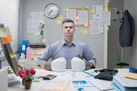 Man in blue shirt showing frustration at a cluttered desk full of paperwork and sticky notes relating to construction accounting software, highlighting the need for an organized solution like Jonas Construction Software. Boxing gloves on hands symbolize the struggle with managing construction related tasks manually.