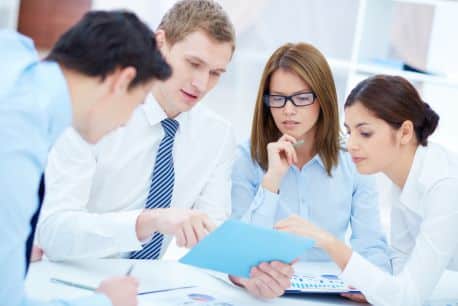 Four specialty contractors, including two male and two female professionals, deeply engrossed in a brainstorming session over key documents. Their collective focus is drawn to a prominent blue folder being held by one of the men. The scene potrays their intensive discussion around the table strewn with papers and a laptop.