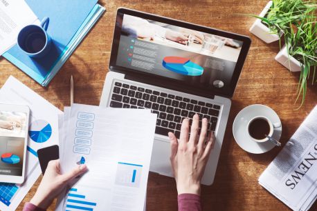 Alt text: A professional analyzing intricate pie-chart data on a laptop using the Jonas Construction Software. Their hands are navigating through a report with a pen, reflecting in-depth interaction with the software. The office desk is filled comprehensively with stacks of documents, a filled coffee cup, and an array of office supplies, conveying an efficient work atmosphere.