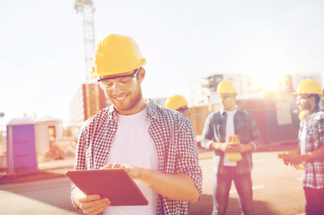 A clean and organized construction site with individuals conversing shows a dedicated engineer focusing on managing tasks using the Jonas Construction Software on his tablet. He is safely dressed in his hardhat, safety glasses, and vests while sporting a cheeky smile as he values the efficiency of handling projects with the software. The software interface on tablet features intuitive dashboard reflecting real-time updates.