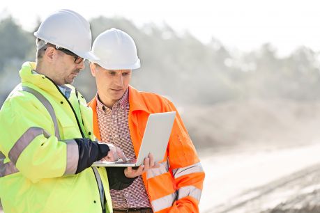 Two construction workers wearing safety attire actively using Jonas Construction Software on a tablet for accounting purposes, with an indistinct backdrop indicating they are at an offsite location.
