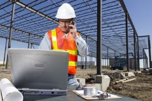 A construction professional in safety gear using Jonas Construction Software on a laptop and discussing project details over the phone at a construction site with steel skeletal structures behind him.