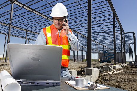 A construction professional in safety gear using Jonas Construction Software on a laptop and discussing project details over the phone at a construction site with steel skeletal structures behind him.