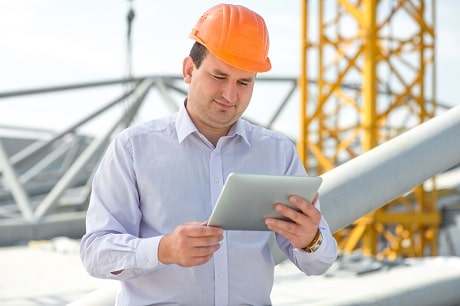 Alt: Construction manager closely examining Jonas Construction Software on a digital tablet at work site, with large industrial structures in the background.