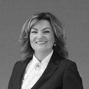 Professional woman with wavy hair smiling at the camera, donning a Jonas Construction branded blouse beneath her suit jacket, alongside subtle earrings. She stands against a neutral, light-toned background.