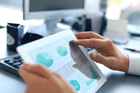 Professional analyzing financial data on a tablet using Jonas Construction Software with partially blurred computer monitor and keyboard in the background, focus is on hand interacting with the tablet.