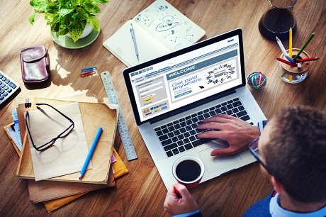 Professional using a laptop displaying a welcome screen for Jonas Construction Software, surrounded by construction related materials such as notebooks, sketches and colored pencils. Additional desk items include glasses, a plant and a cup of coffee.