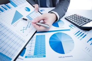 A business professional meticulously reviewing financial reports, filled with detailed charts and graphs specific to the construction industry. He's thoughtfully holding a pen, calculating figures on a nearby calculator, ensuring data accuracy for specialty contractors. The scene depicts an important part of job project management in a formal office environment.