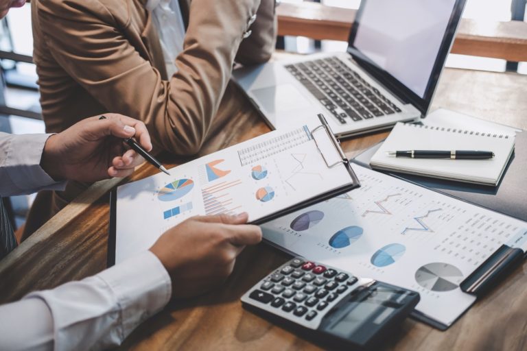 Two specialty contractors discussing data on a chart, surrounded by various work items such as laptops, documents, calculator and a notebook at a wooden table. One of them is actively pointing towards the chart to facilitate collaboration.