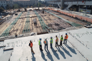 Eight construction workers in safety gear having a discussion at a large construction site, using Jonas Construction Software for planning. Aerial view of the site showcases extensive steel reinforcements prepared for concrete placement.