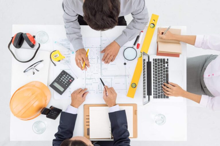 Three professionals, consisting of two men and one woman, having an in-depth conversation about Jonas Construction architectural plans on a white table. The environment is adorned with essential work tools such as drafting instruments and a laptop. A construction safety helmet is also present, symbolizing safety protocol adherence within the industry.