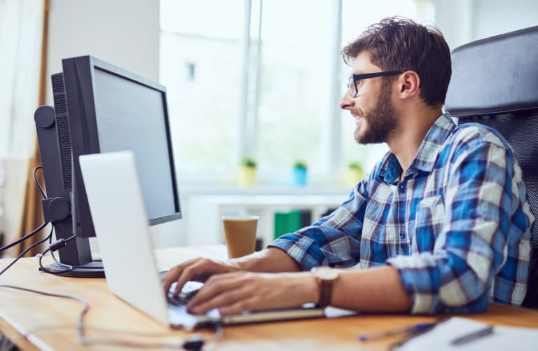 Alt text: A bespectacled-bearded man in a plaid shirt using Jonas Construction Software on a dual monitor setup in a well-lit office, with an accompanying coffee cup.