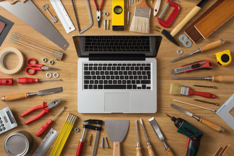 Alt text: A laptop displaying Jonas Construction Software, strategically positioned amidst an array of construction tools such as a hammer, screwdrivers, paintbrush, measuring tape, screws and woodworking tools on a wooden work desk - symbolizing the integration of technology in streamlining construction processes.