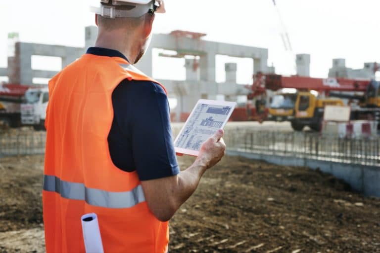 A construction professional analyzing blueprints on a digital tablet, with heavy machineries and bustling site activity visible in the backdrop, demonstrating the use of Jonas Construction software for straightened productivity.