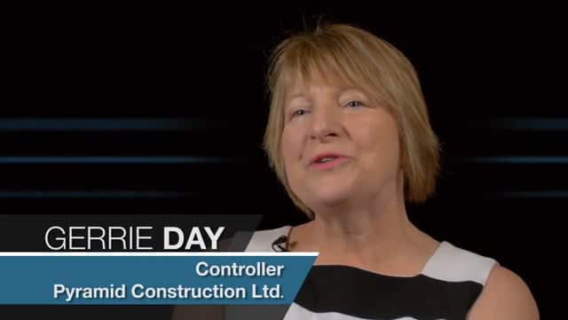 in a transparent box at the bottom of the screen. The background shows an office environment with sleek white desks and abstract wall art.
Alt-text: Professional woman Gerrie Day, Controller at Jonas Construction Ltd., being interviewed in a modern office setting.