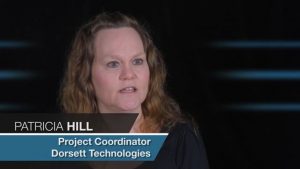 Alt text: "Medium-length brown haired woman, Patricia Hill, talking in an interview setting with her title 'Project Coordinator at Jonas Construction' written below. The background is dimly lit for emphasis on her.