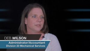Alt Text: "A professional woman named Deb Wilson, who is an administrator/accountant for Division 23 Mechanical Contractors, speaking attentively in an interview. The blue-toned background artfully blurred to focus on her.