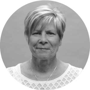 Alt text: Smiling middle-aged woman with short hair wearing a white lacy blouse and necklace featuring Jonas Construction logo, against a plain grey background.