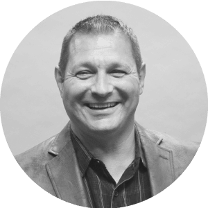 Portrait of a cheerful middle-aged Caucasian man with short hair, wearing a jacket over a shirt featuring the Jonas Construction logo. His image is styled in monochrome and neatly cropped within a circular frame.