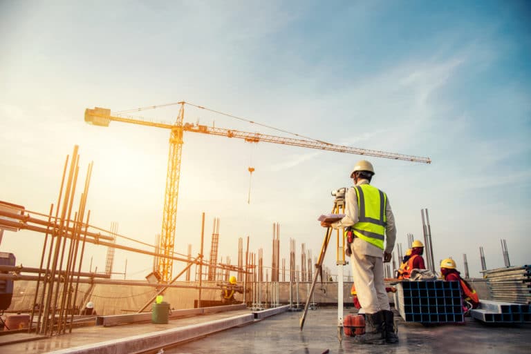 Image of construction workers in safety outfits performing various tasks at a worksite with a towering crane against an evening sky. One worker is diligently examining plans on clipboard amidst steel beams and scaffolding.