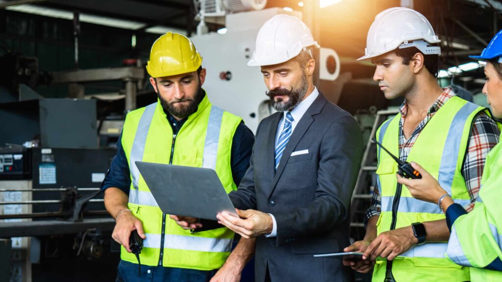 Four construction professionals in safety gear analyzing project details on a laptop displaying Jonas Construction Software, with an industrial backdrop suggestive of on-site working conditions.