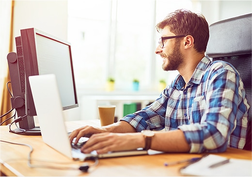 Alt text: A man with beard and glasses, dressed in a plaid shirt, diligently working on a dual monitor setup. His fingers swiftly hitting keys on the keyboard while a cup of coffee sits nearby. A window with warm sunlight filters in, illuminating the workspace.