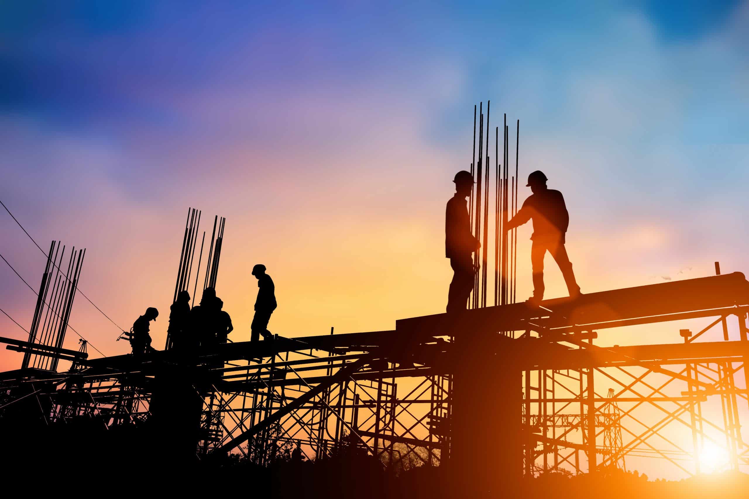 Silhouettes of specialty contractors working on scaffolding amid protruding steel rods at a construction site, set against a dramatic sunset sky.