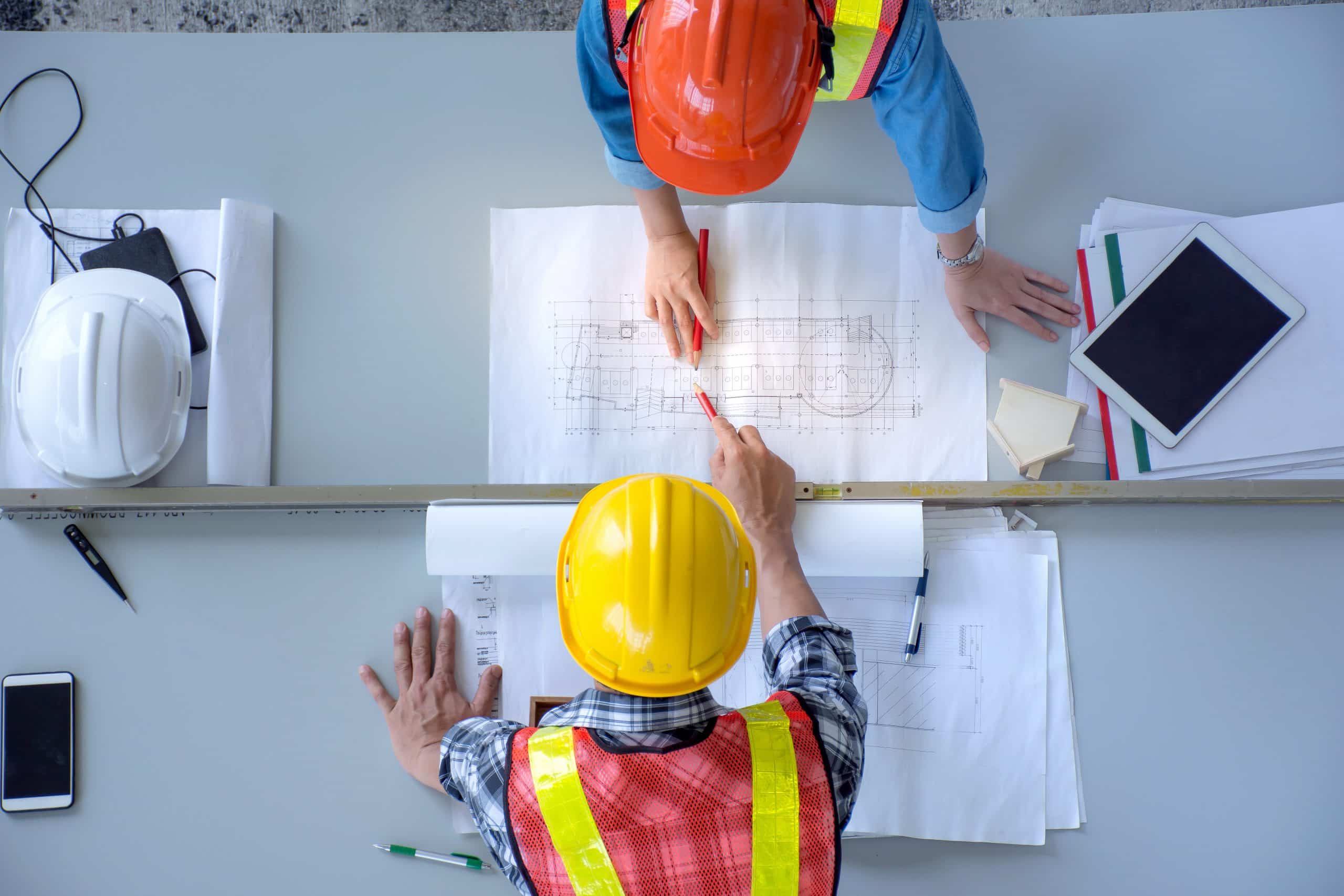 Two professionals outfitted in safety gear engrossed in a discussion over architectural plans. They are surrounded by essential tools of their trade, including a laptop running Jonas Construction Software and a smartphone. Additional hard hats can be seen, signifying the level of safety they practice. A bird's eye perspective on the scene.