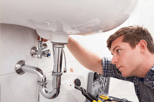 Alt text: "A specialty contractor in casual attire intently working on sink pipe repairs using a wrench, with various tools scattered nearby in a neat and clean tiled bathroom.