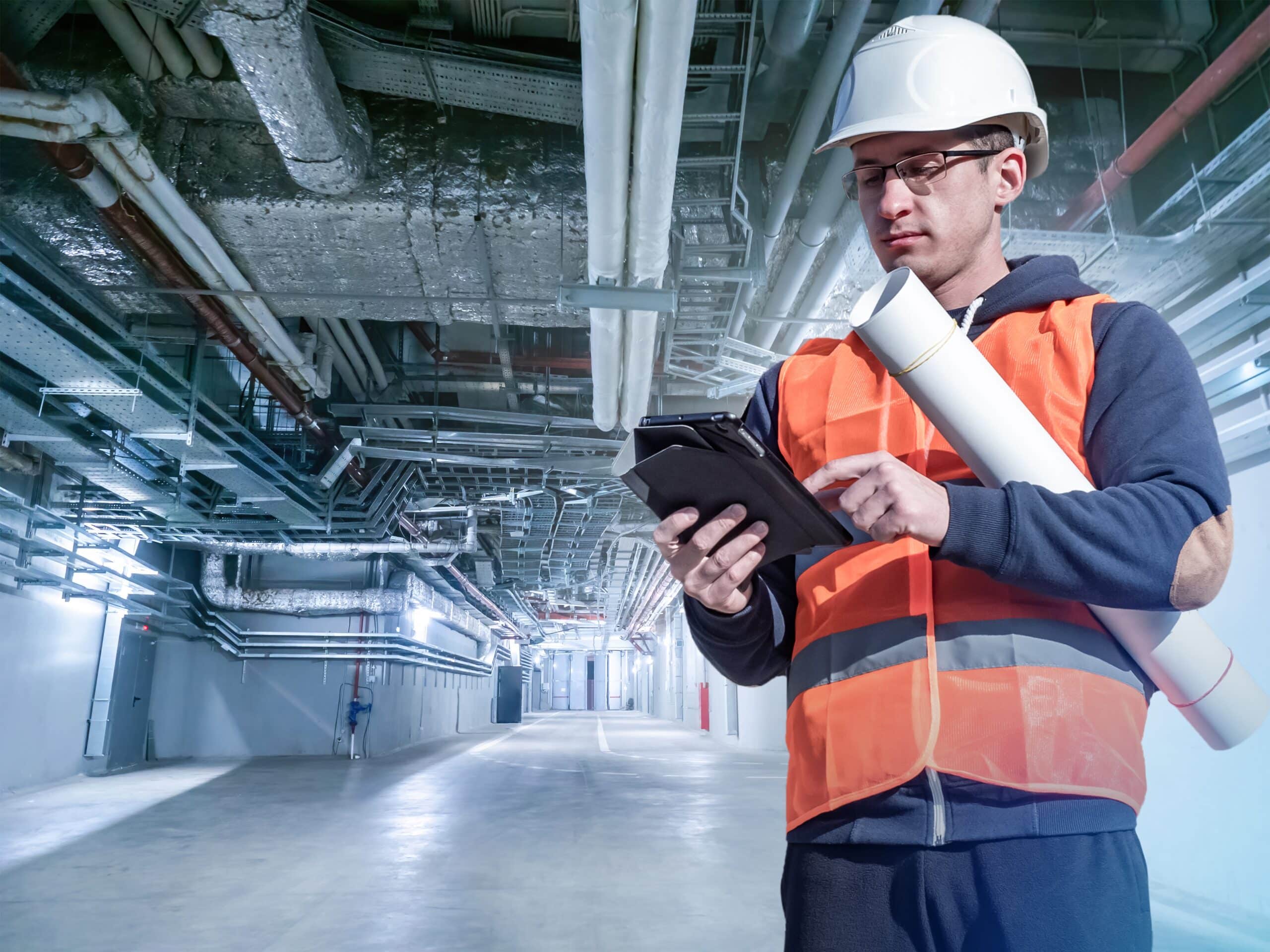 A construction worker in safety attire using Jonas Construction Software on a digital tablet, holding rolled blueprints in an industrial basement with overhead pipes.