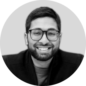 Alt Text: "Grayscale portrait of a cheerful money wearing glasses and a dark jacket, with short black hair and beard against a soft, light gray background.