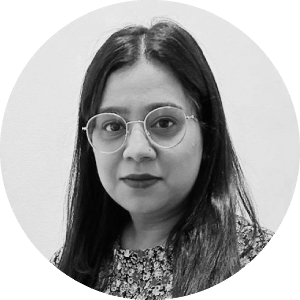 Portrait of a calm South Asian woman with shoulder-length hair and round glasses, wearing a floral top against a plain background.