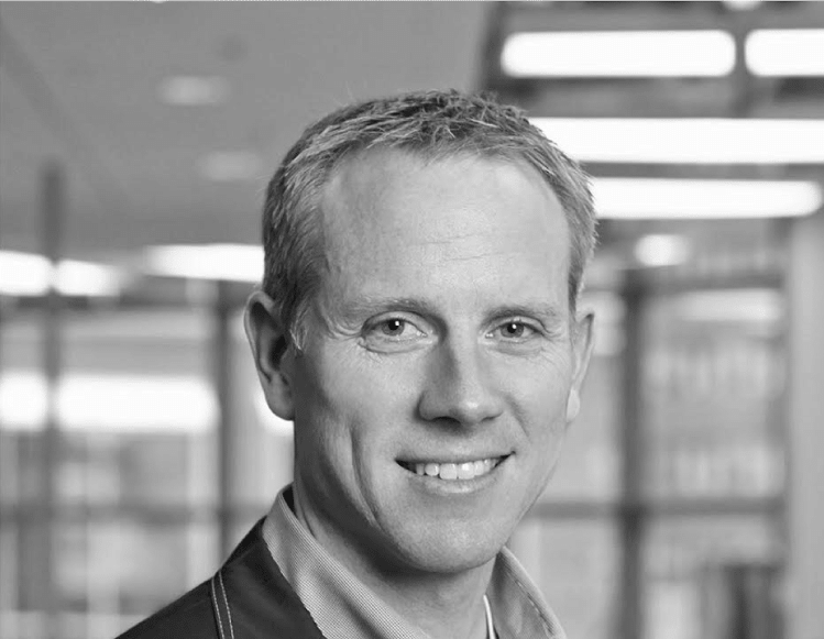 Middle-aged man with short hair, smiling and looking directly at the camera. He's wearing a casual jacket and standing indoors against a blurred office background featuring Jonas Construction Software signage.